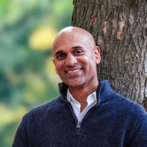 Professional headshot of smiling Sportball employee in navy pullover against tree and blurred greenery